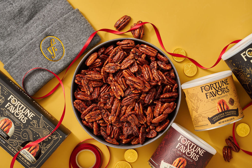 A bowl of Fortune Favors candied pecans with cans, a knit hat and ribbon laid out for gifting